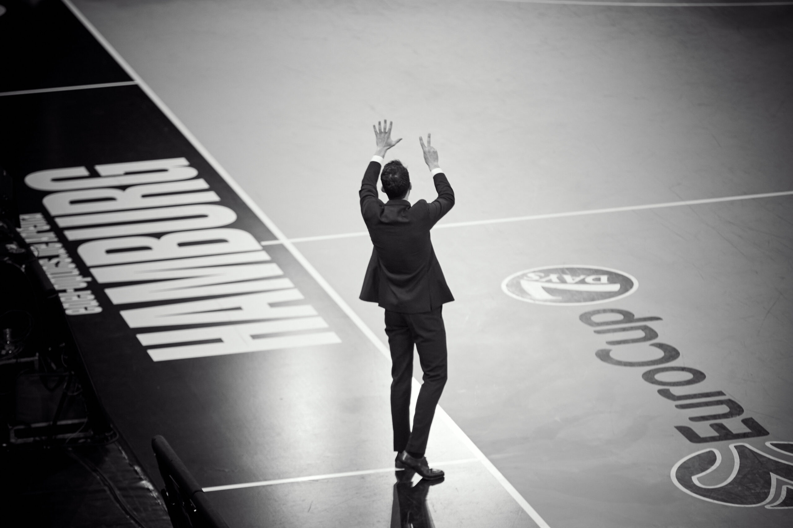 Mann in schwarzem Anzug steht an der Seitenlinie eines Basketballfeldes und hält sieben Finger hoch