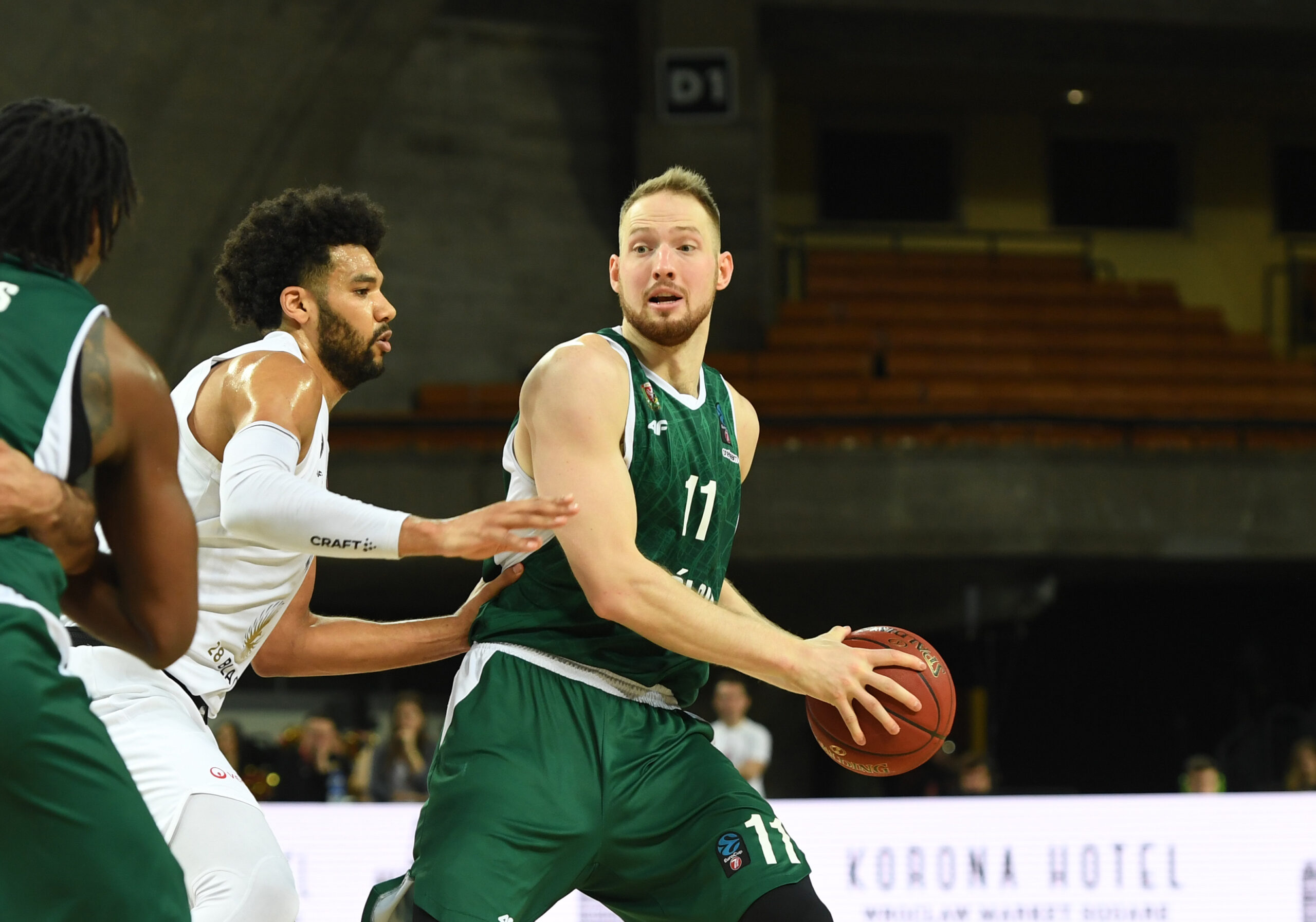 Spieler in grüner Uniform hält einen Basketball in den Händen, Spieler in weißer Uniform verteidigt ihn aktiv Im Hintergrund leere Zuschauerränge
