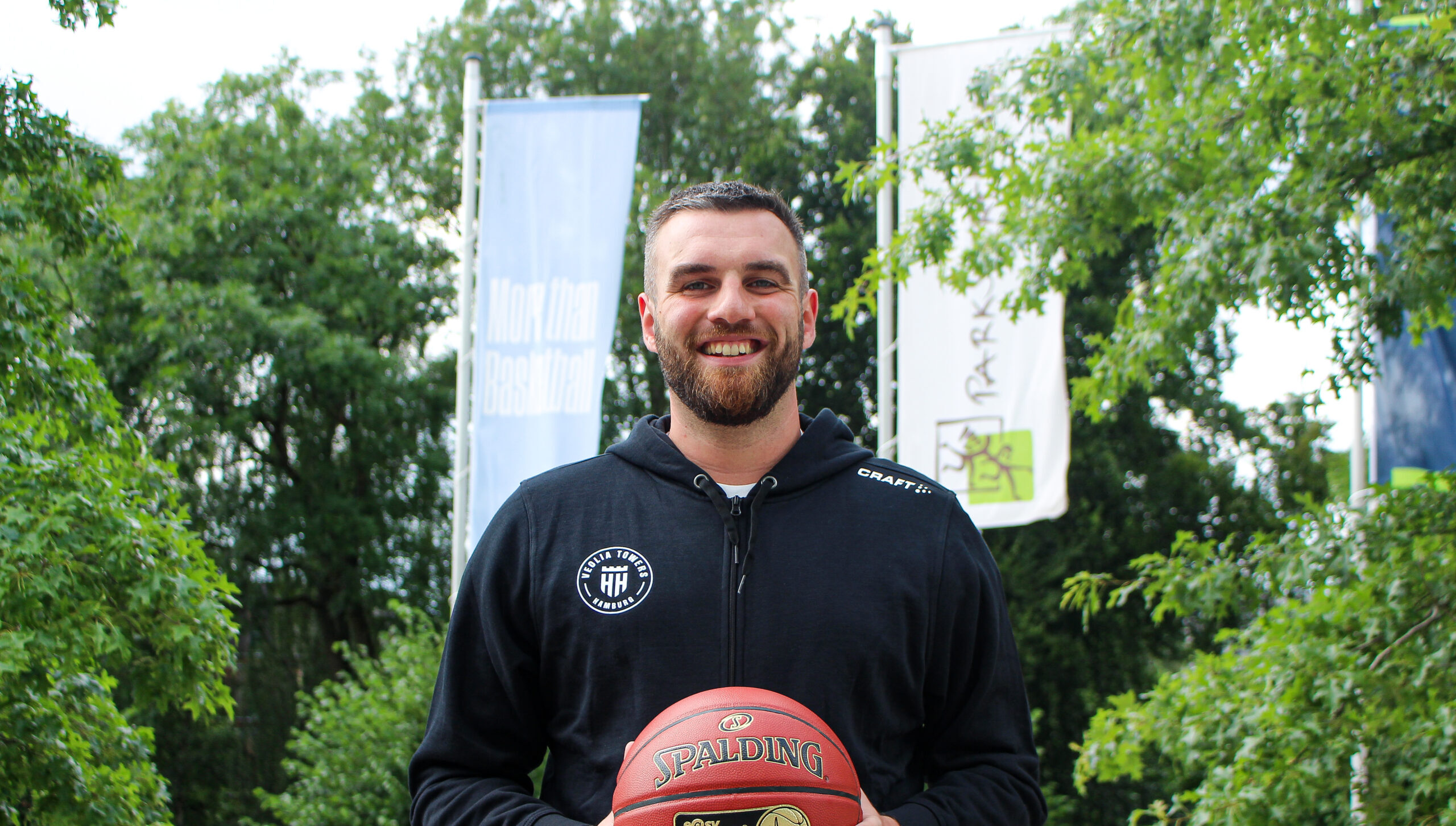 Person trägt schwarzen Kapuzenpullover und hält einen orangefarbenen Spalding-Basketball vor grünem Hintergrund