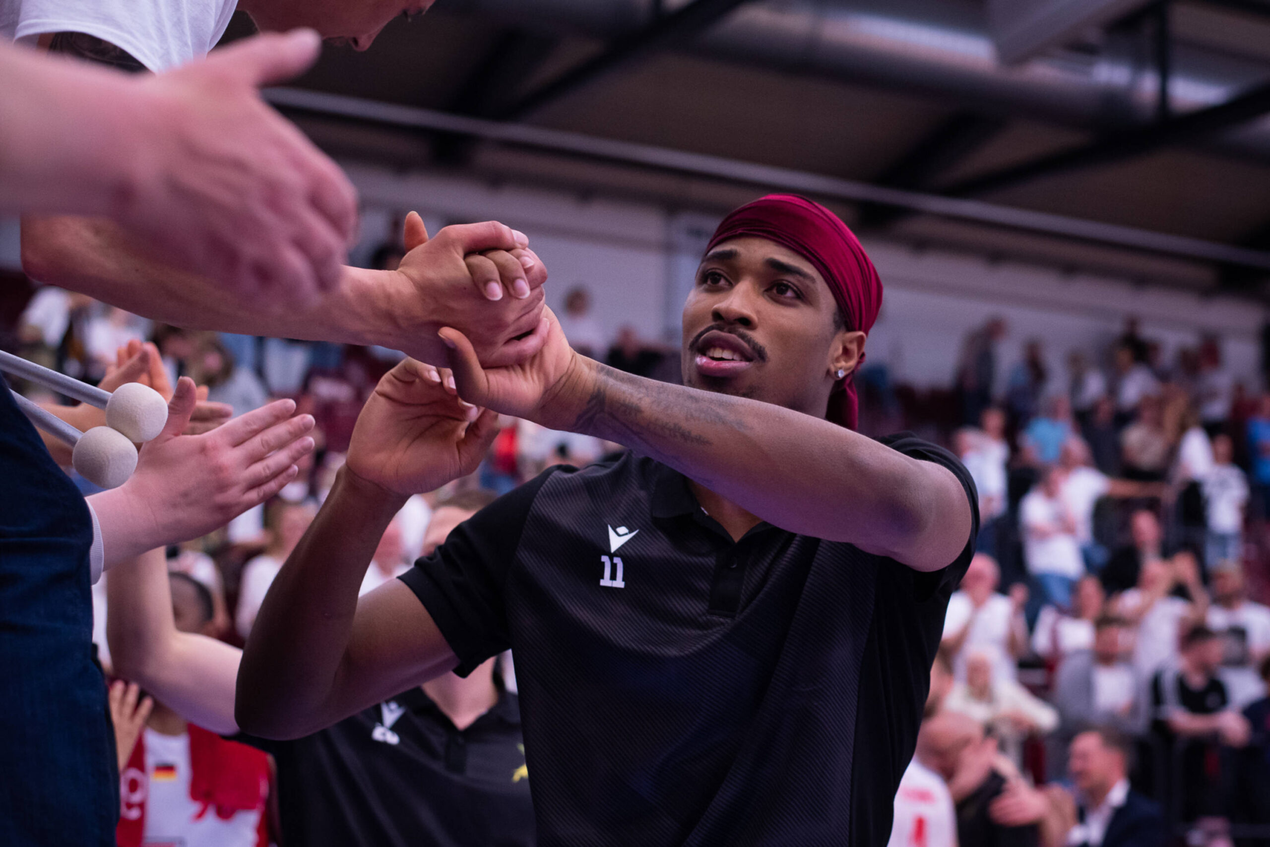 Mann in schwarzem Shirt mit rotem Kopftuch klatscht bei Fans ab. Im Hintergrund Zuschauer