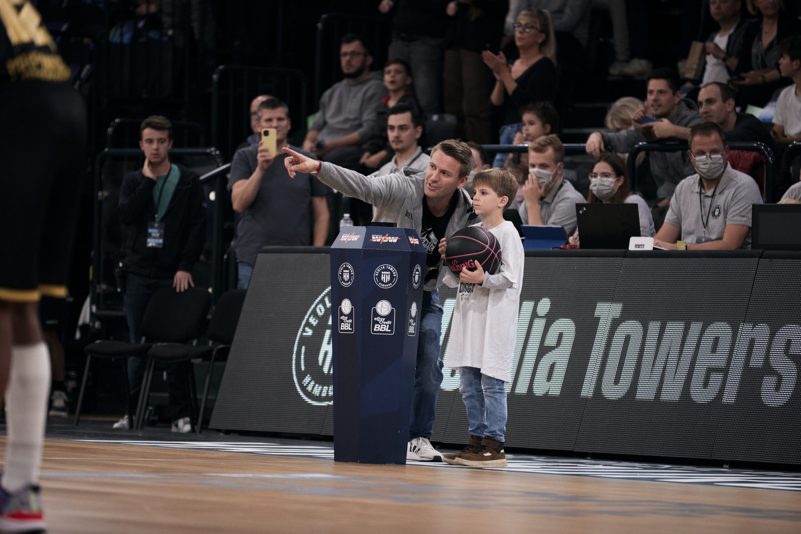 Ein Junge hält einen Basketball und steht neben einem Mann, der auf das Spielfeld zeigt, während Zuschauer im Hintergrund applaudieren.