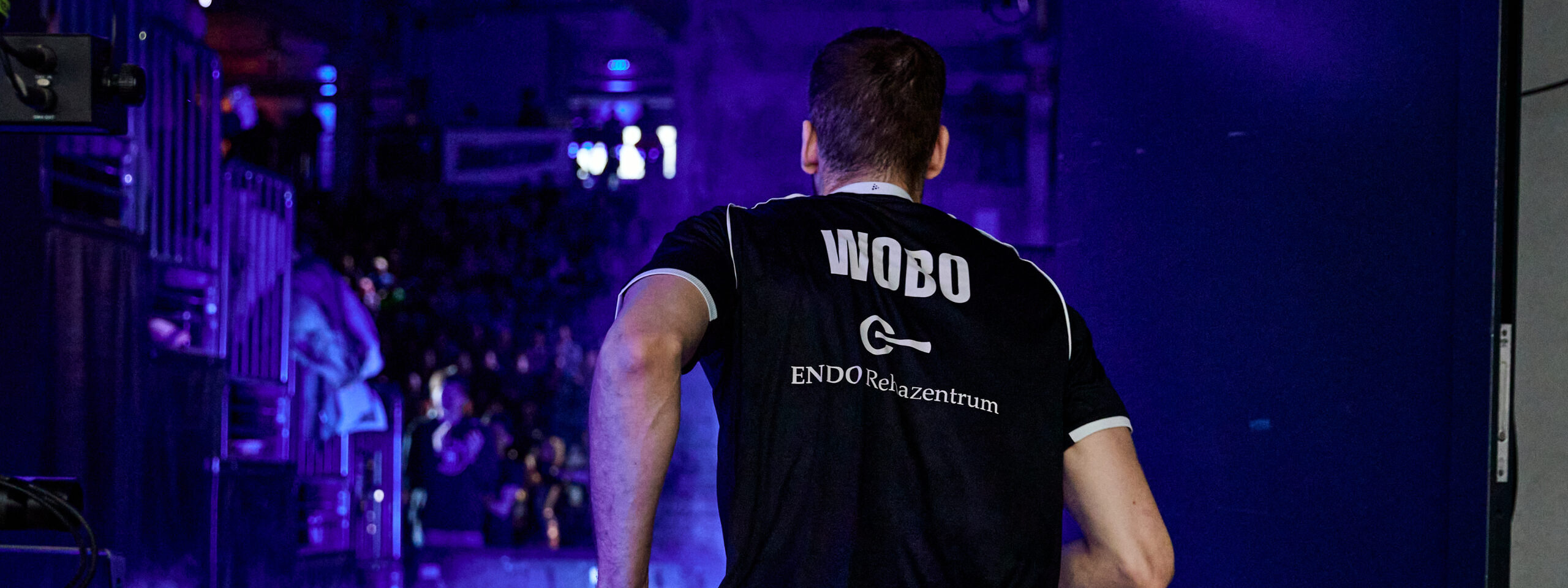 Basketballspieler Dennis Fischer von hinten in schwarzem Trikot mit Aufschrift 'WOBO' und 'ENDO Relaxzentrum' beim Betreten des Spielfelds