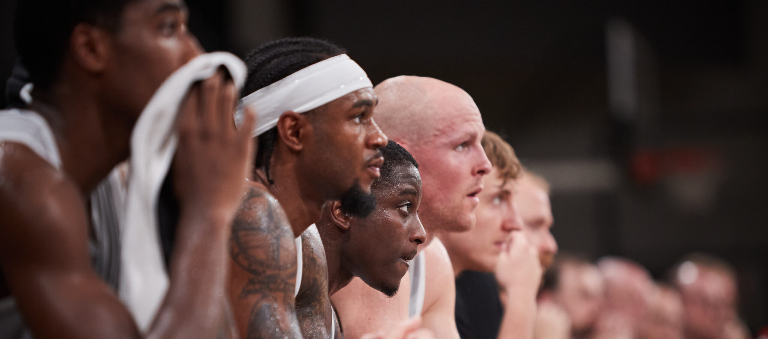Mehrere Basketballspieler sitzen nebeneinander auf der Bank, tragen weiße Trikots und beobachten das Spiel konzentriert.