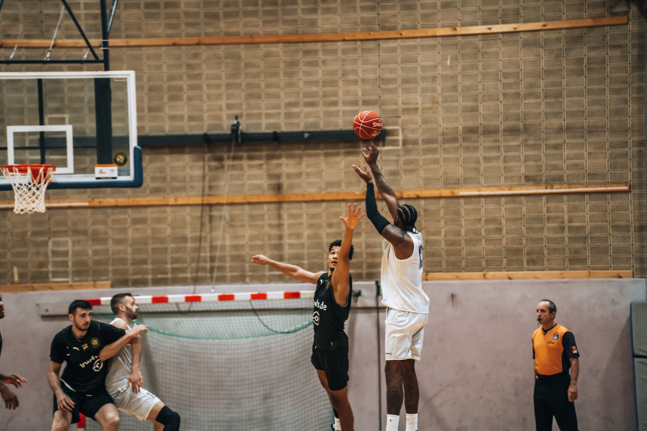 Basketballspieler in weißem Trikot springt zum Wurf, während ein Spieler in schwarzem Trikot versucht zu blocken, Hallenwand und Korb im Hintergrund