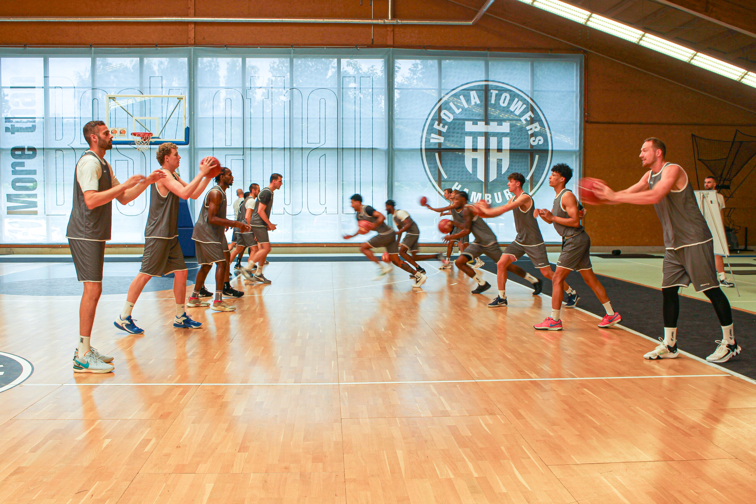 Basketballspieler in schwarzen Trikots beim Passspiel auf einem Hallenparkett mit Logo der Hamburg Towers im Hintergrund