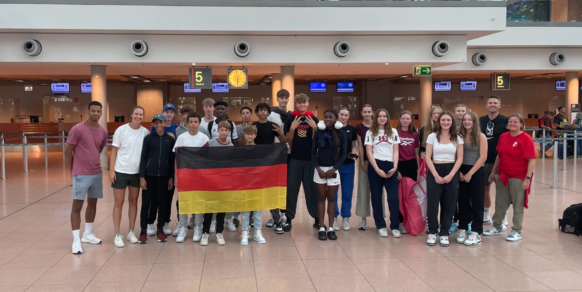 Gruppe von Jugendlichen und jungen Erwachsenen steht in einem Flughafen-Terminal vor Check-in-Schaltern, zwei Personen halten eine deutsche Flagge