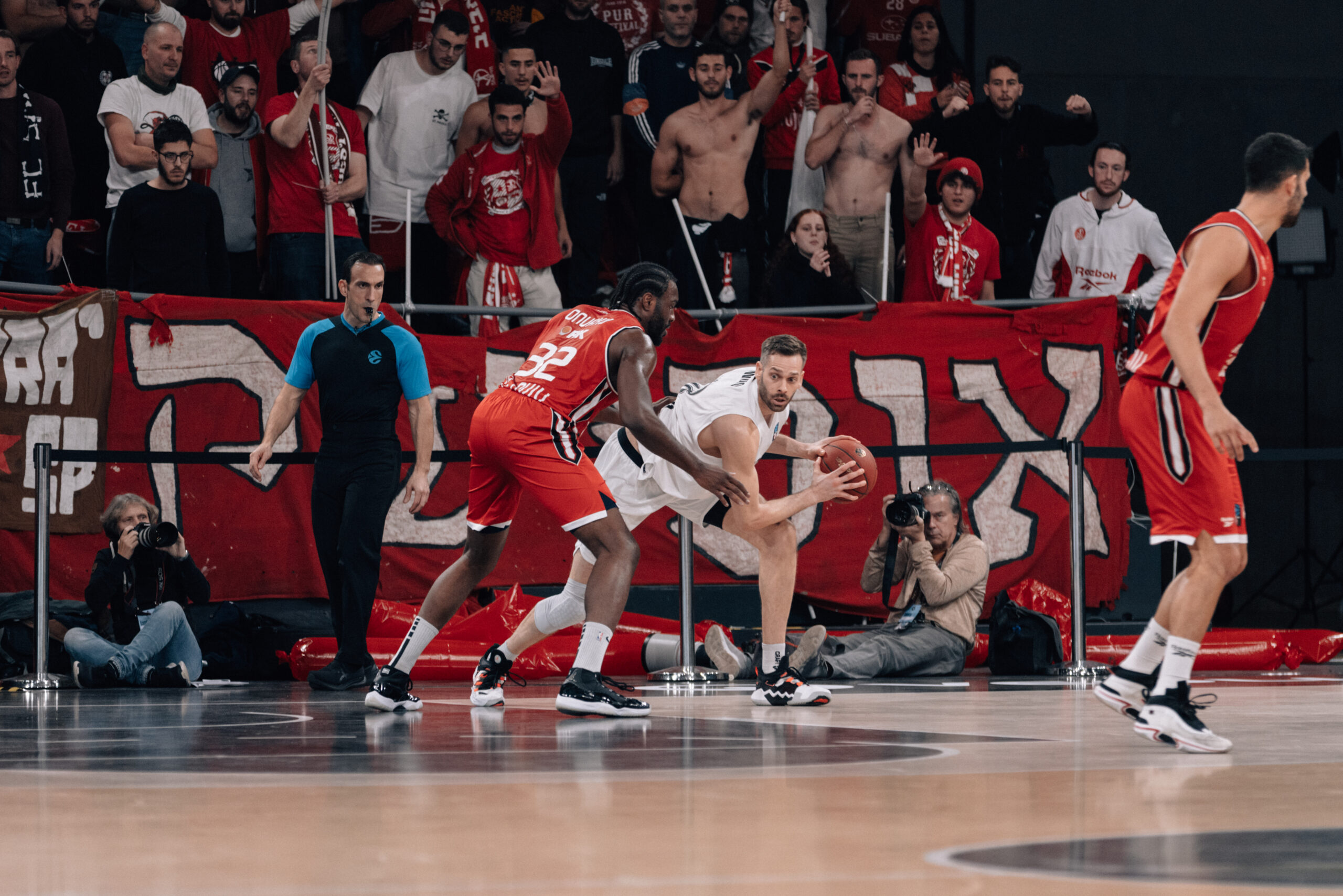 Basketballspieler in weißer und roter Mannschaftskleidung kämpfen um den Ball auf dem Spielfeld, Zuschauer im Hintergrund