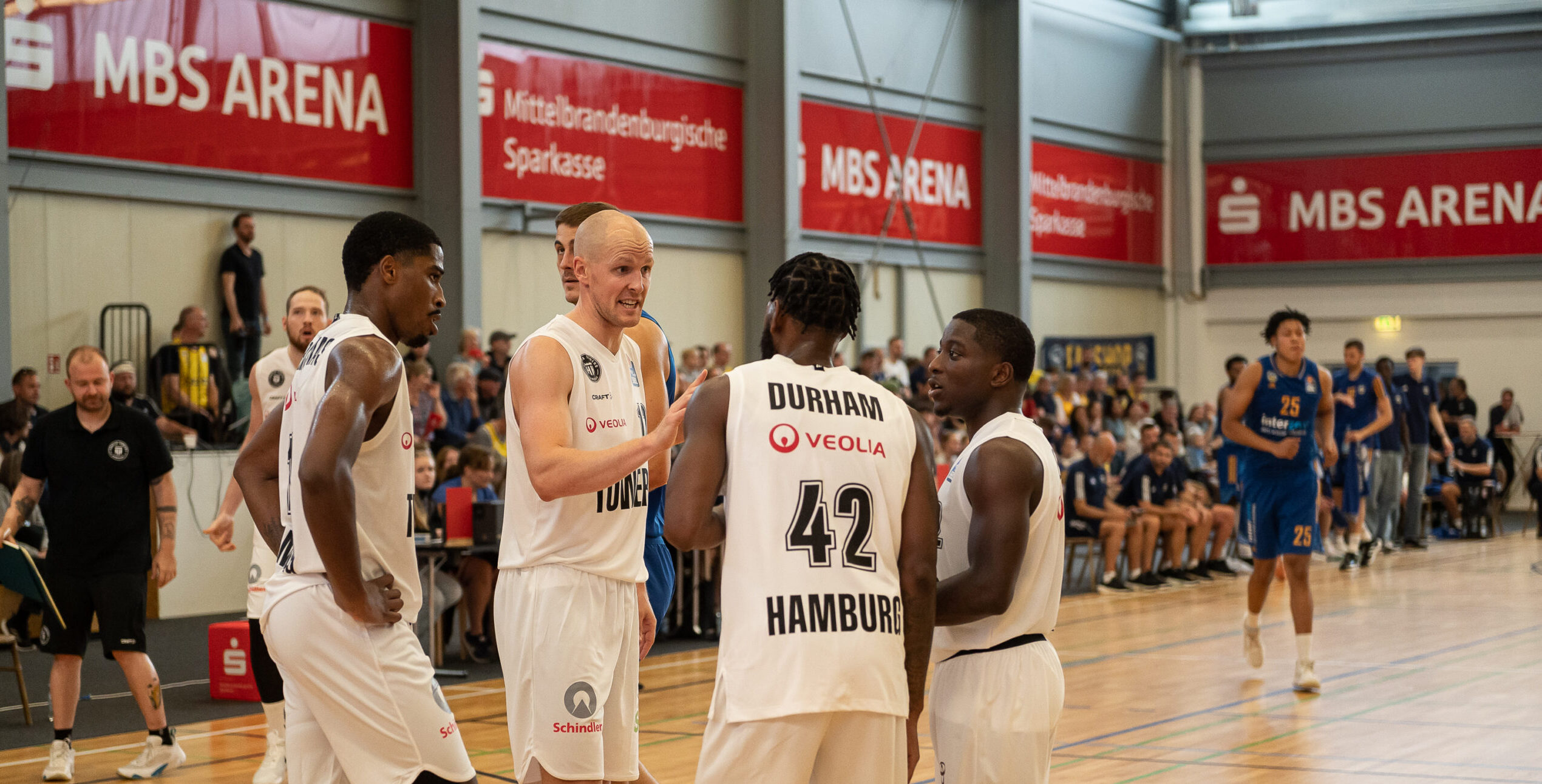 Vier Basketballspieler in weißen Trikots mit der Aufschrift 'Durham 42 Hamburg' stehen in einer Sporthalle mit Zuschauern im Hintergrund.