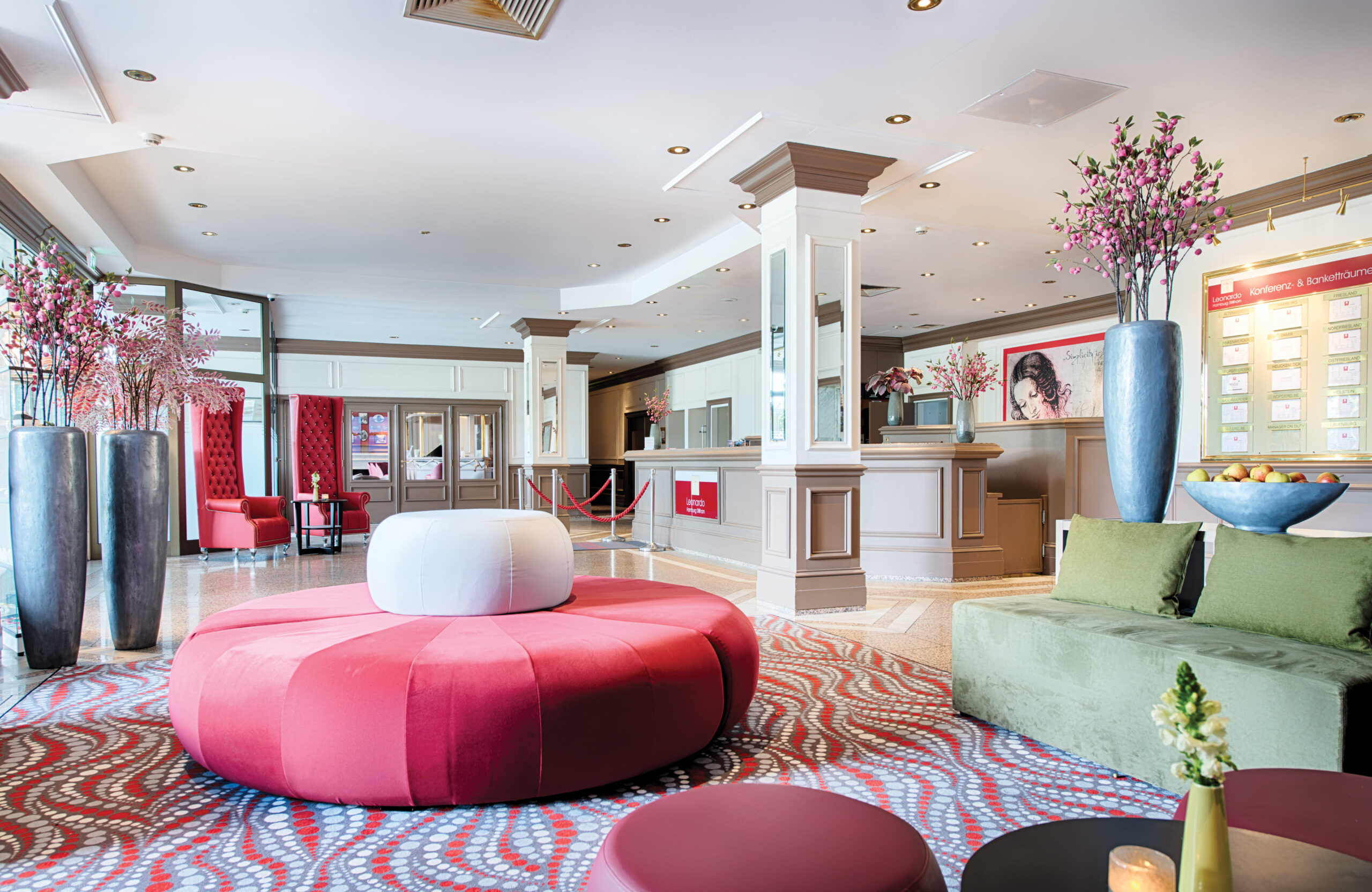 Hotel-Lobby mit rundem roten Sitzmöbel, grünem Sofa, Rezeptionstresen und großen Vasen mit rosa Blumen