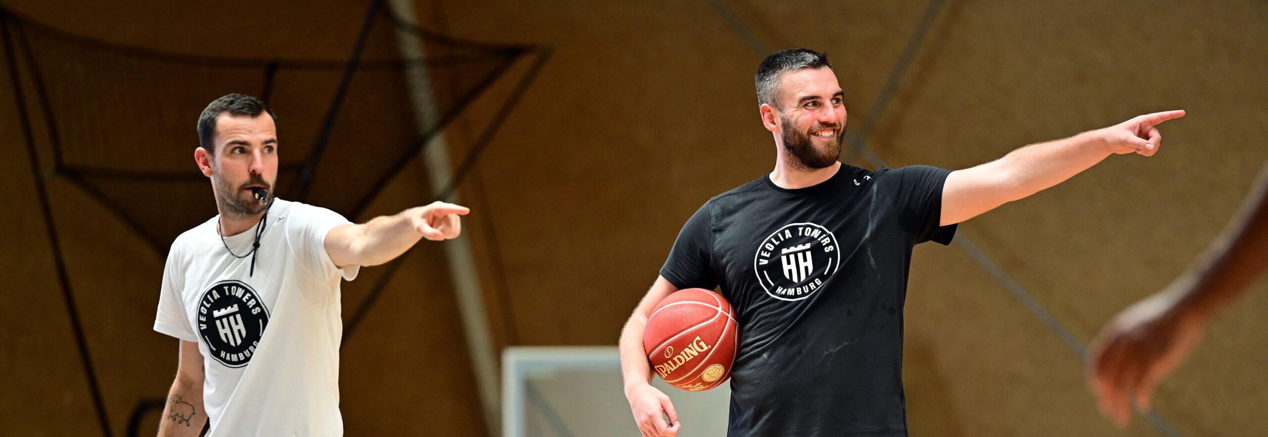 Zwei Männer in Sportkleidung zeigen mit ausgestrecktem Arm in dieselbe Richtung, einer hält einen Basketball