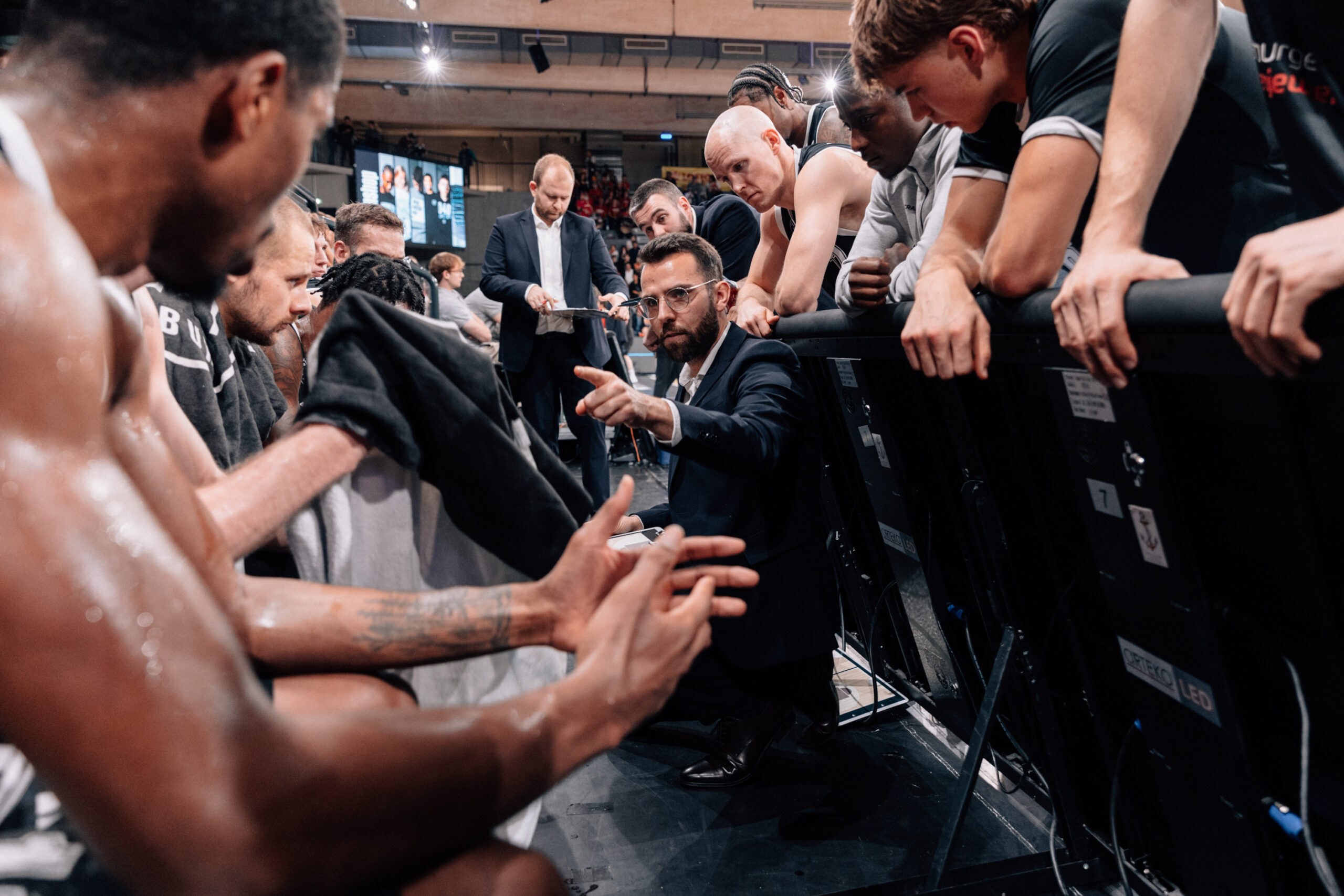 Basketballtrainer gibt Anweisungen an Spieler während einer Auszeit, Spieler tragen schwarze Trikots und sitzen auf der Bank
