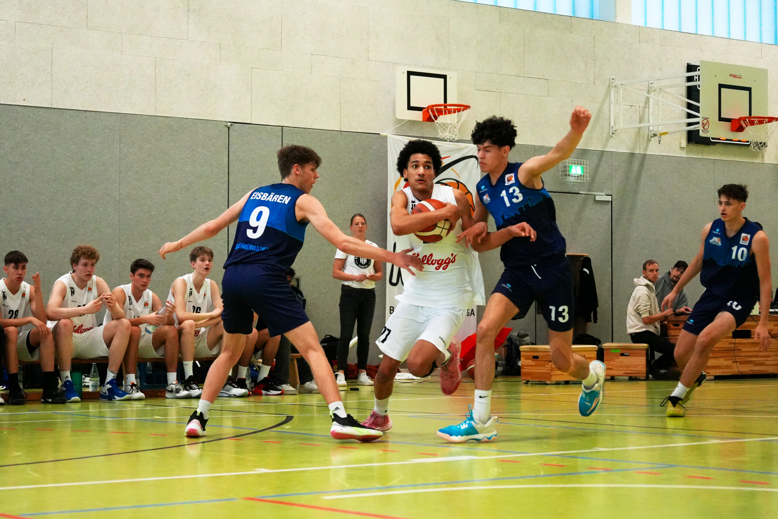 Jugendliche Basketballspieler in einem Hallenspiel, zwei Spieler in blauen Trikots verteidigen gegen einen Spieler in weißem Trikot mit Ball