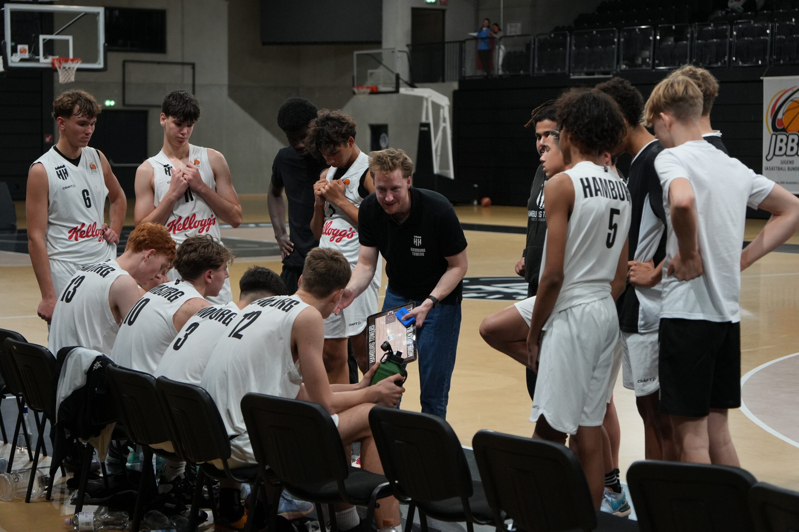 Basketballtrainer gibt Anweisungen an sein Team während einer Auszeit in der Halle, Spieler tragen weiße Trikots mit 'Kellogg's' und 'Hamburg'