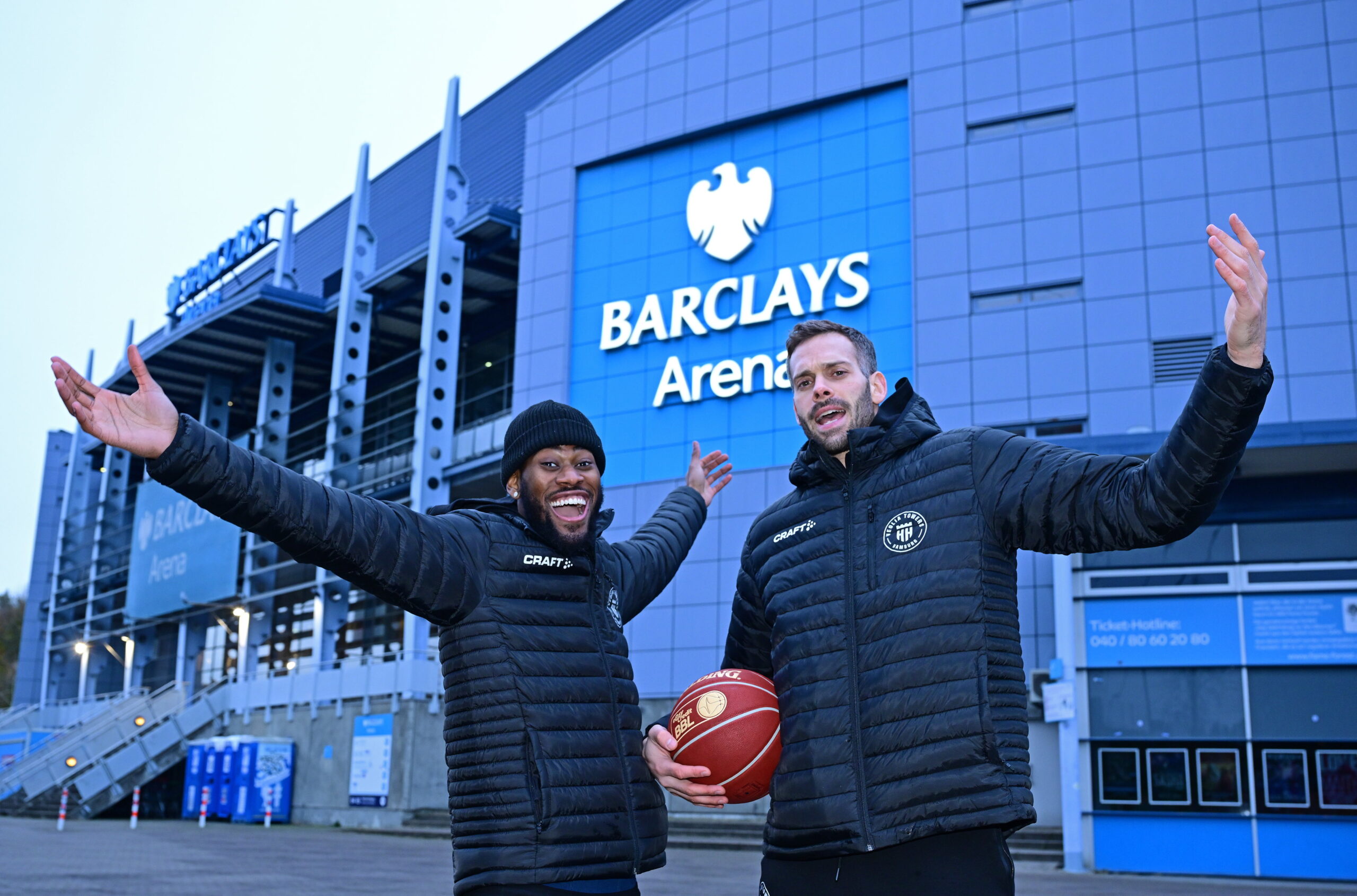 Zwei Männer in schwarzen Jacken vor der Barclays Arena, einer hält einen Basketball, beide heben die Arme