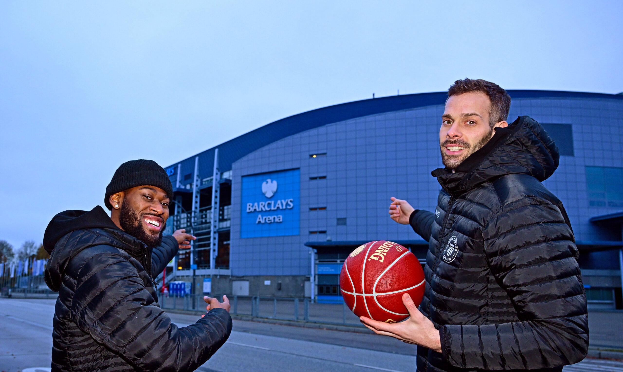 Zwei Männer in schwarzen Jacken vor der Barclays Arena, einer hält einen Basketball, beide heben die Arme