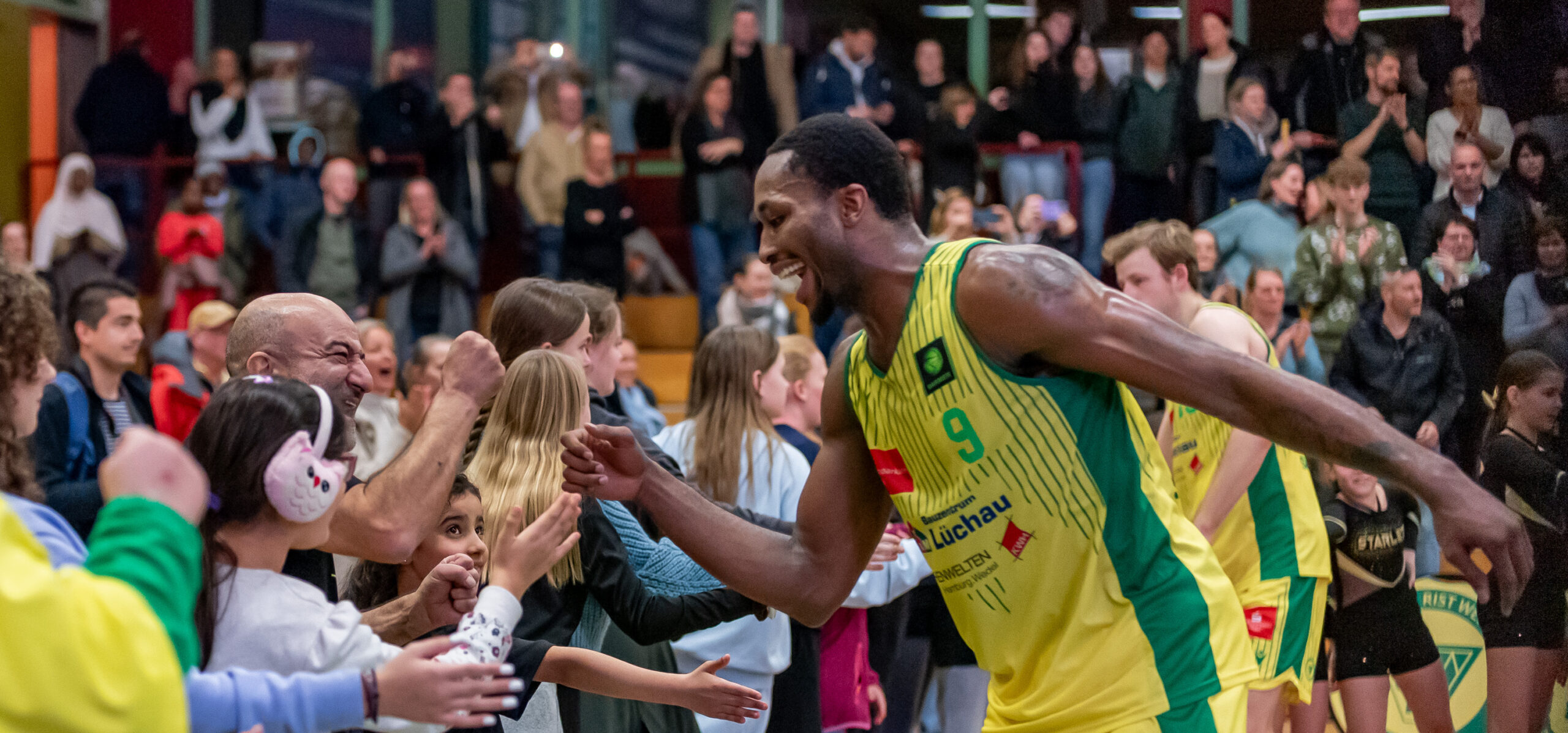 Basketballspieler in gelb-grüner Uniform mit der Nummer 9 gibt Fans nach einem Spiel High-Fives in einer Halle mit Zuschauern