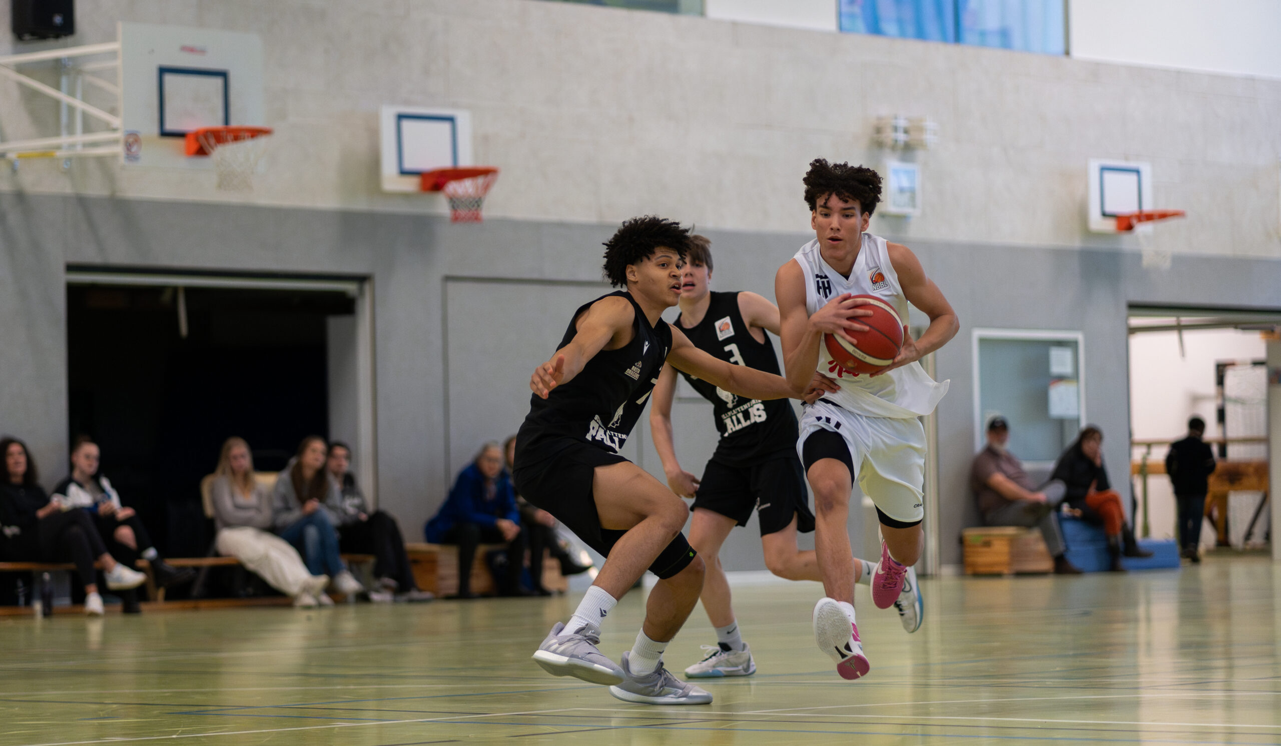 Junger Basketballspieler in weißem Trikot dribbelt den Ball, zwei Gegner in schwarzen Trikots verteidigen in einer Halle
