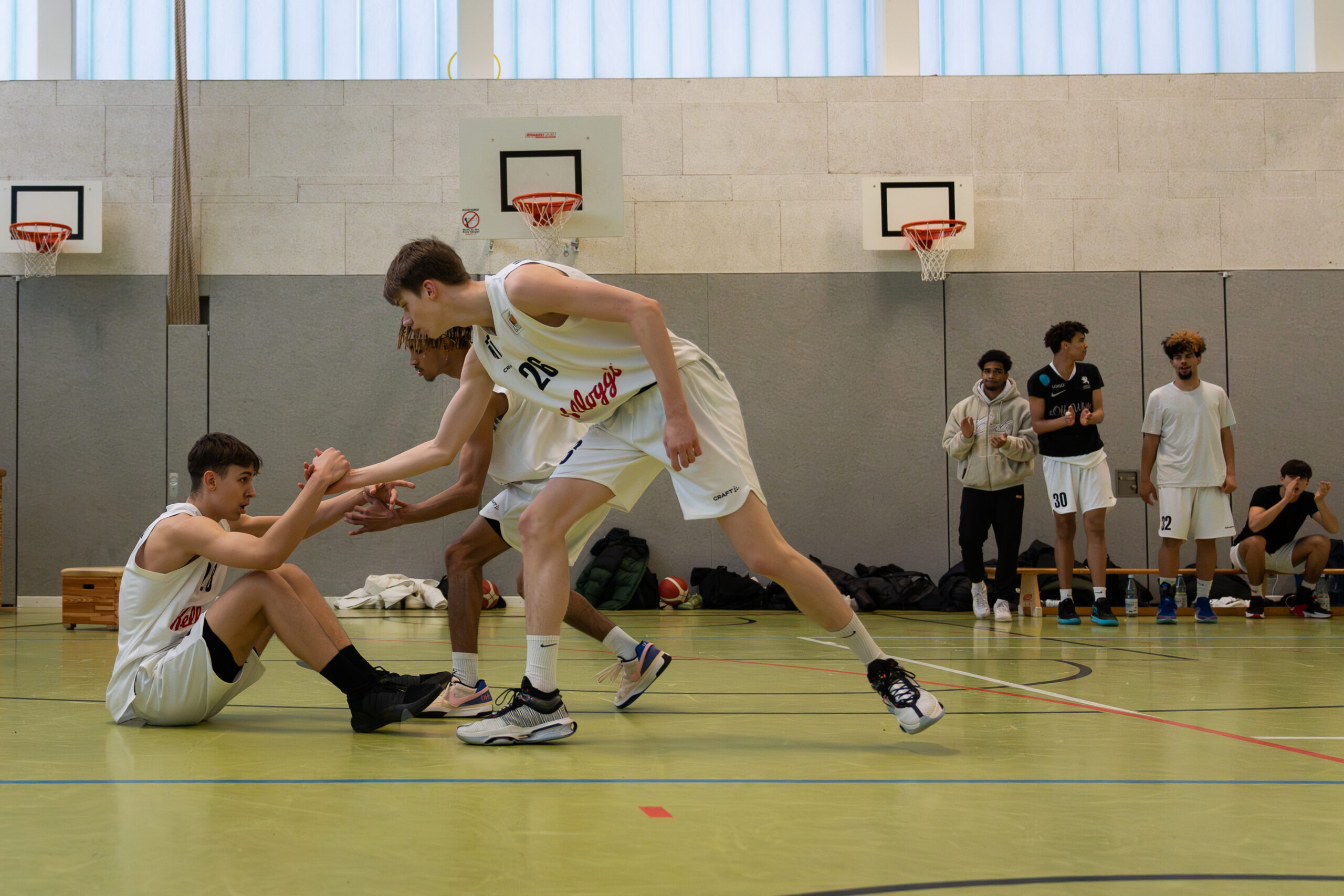 Zwei Basketballspieler in weißen Trikots helfen einem am Boden sitzenden Spieler hoch, im Hintergrund Halle mit Basketballkörben und junge Männer vor einer Bank die klatschen