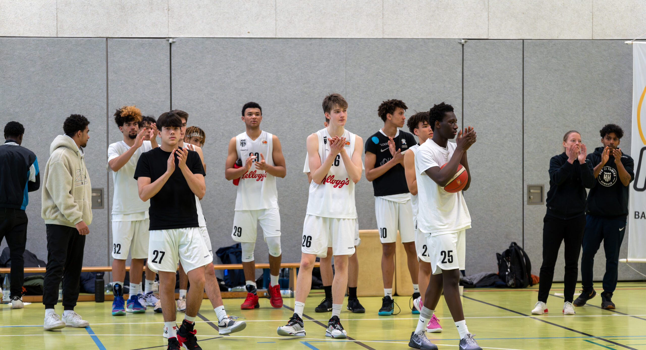 Basketballteam in weißen Trikots steht auf dem Spielfeld und klatscht Richtung Zuschauer