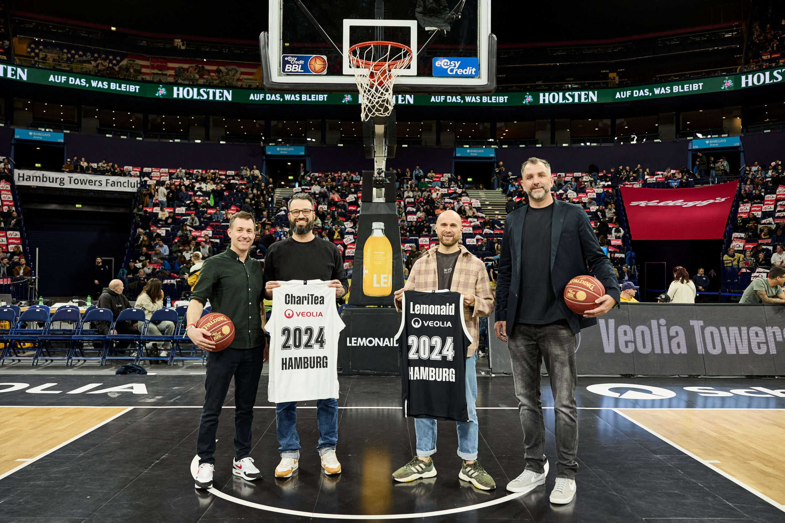 Vier Männer stehen auf einem Basketballfeld in Hamburg, zwei halten Basketbälle, zwei tragen Trikots mit '2024 Hamburg' und Sponsorennamen