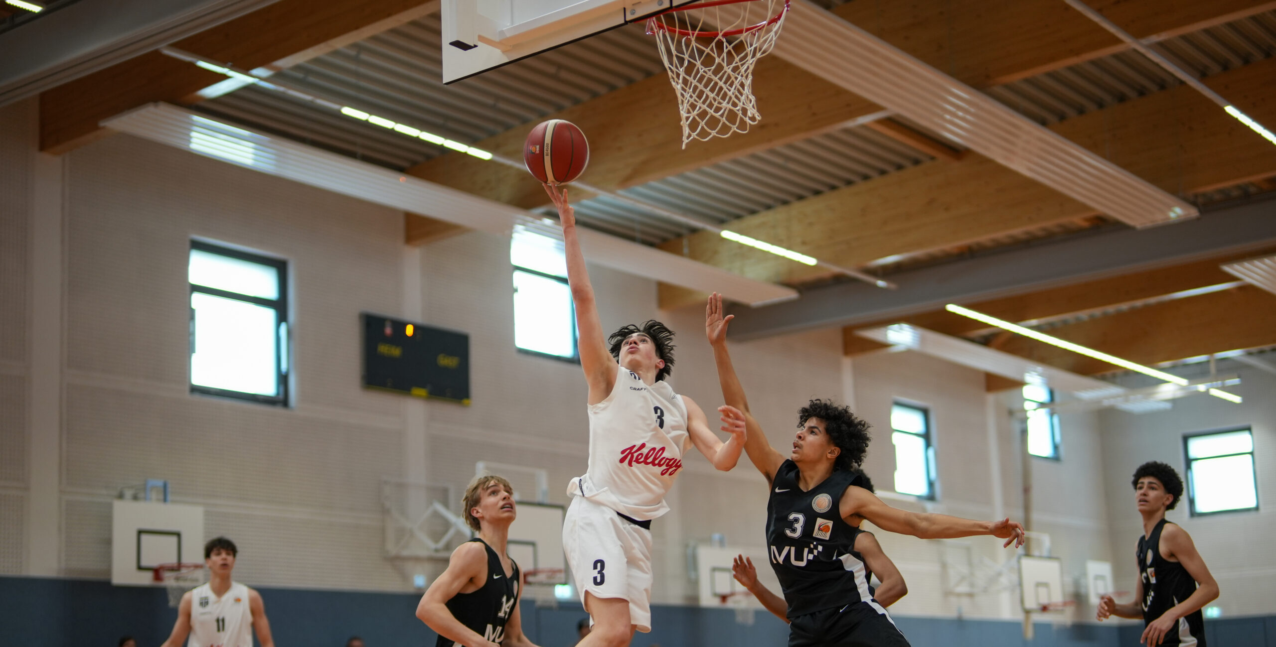 Basketballspieler in weißem Trikot mit der Nummer 3 wirft Korbleger gegen Verteidiger in schwarzem Trikot mit der Nummer 3 in einer Halle