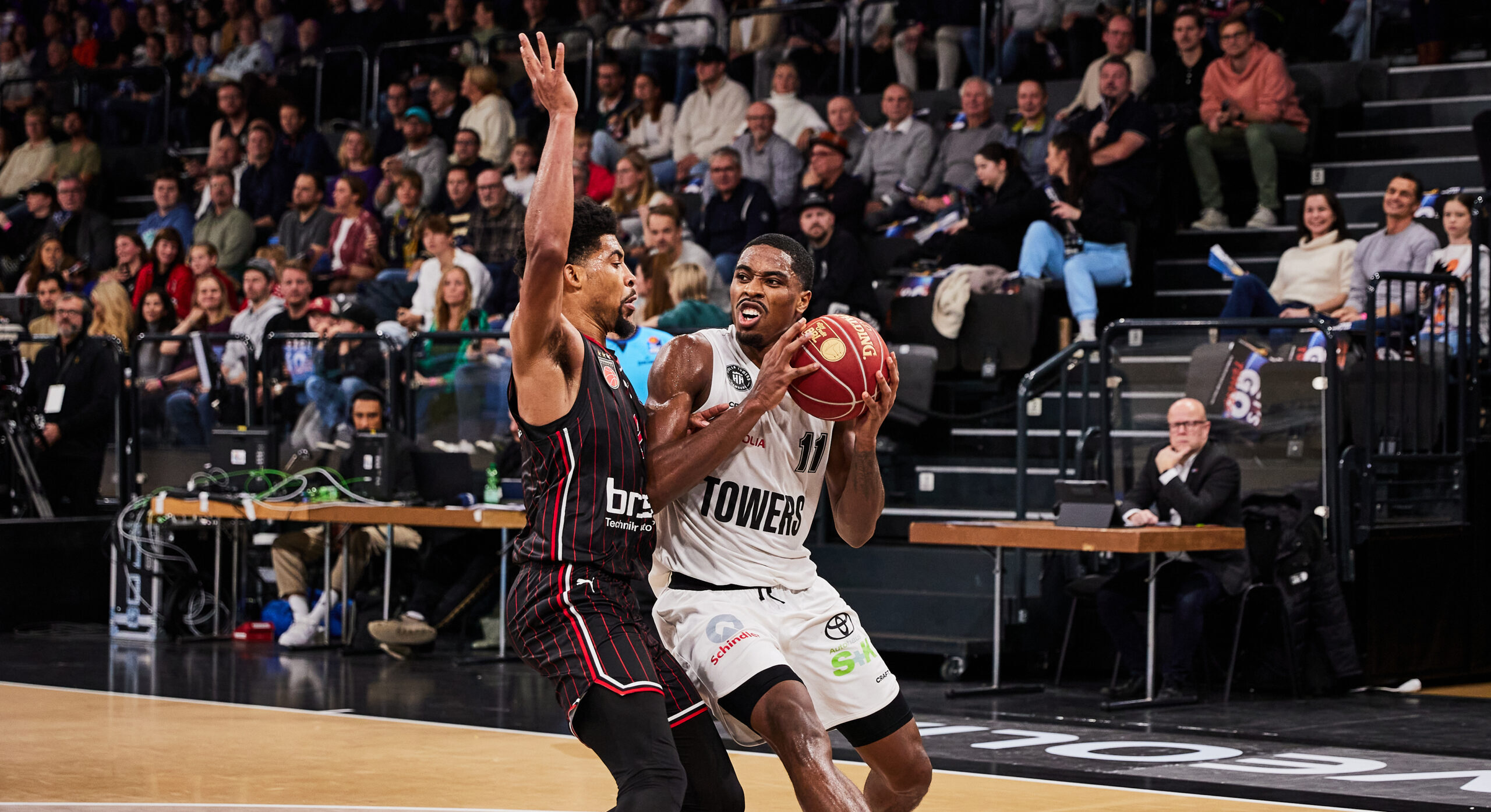 Basketballspieler der Towers mit Ball in der Hand, verteidigt von einem Spieler des Gegners in schwarzem Trikot, Zuschauer im Hintergrund