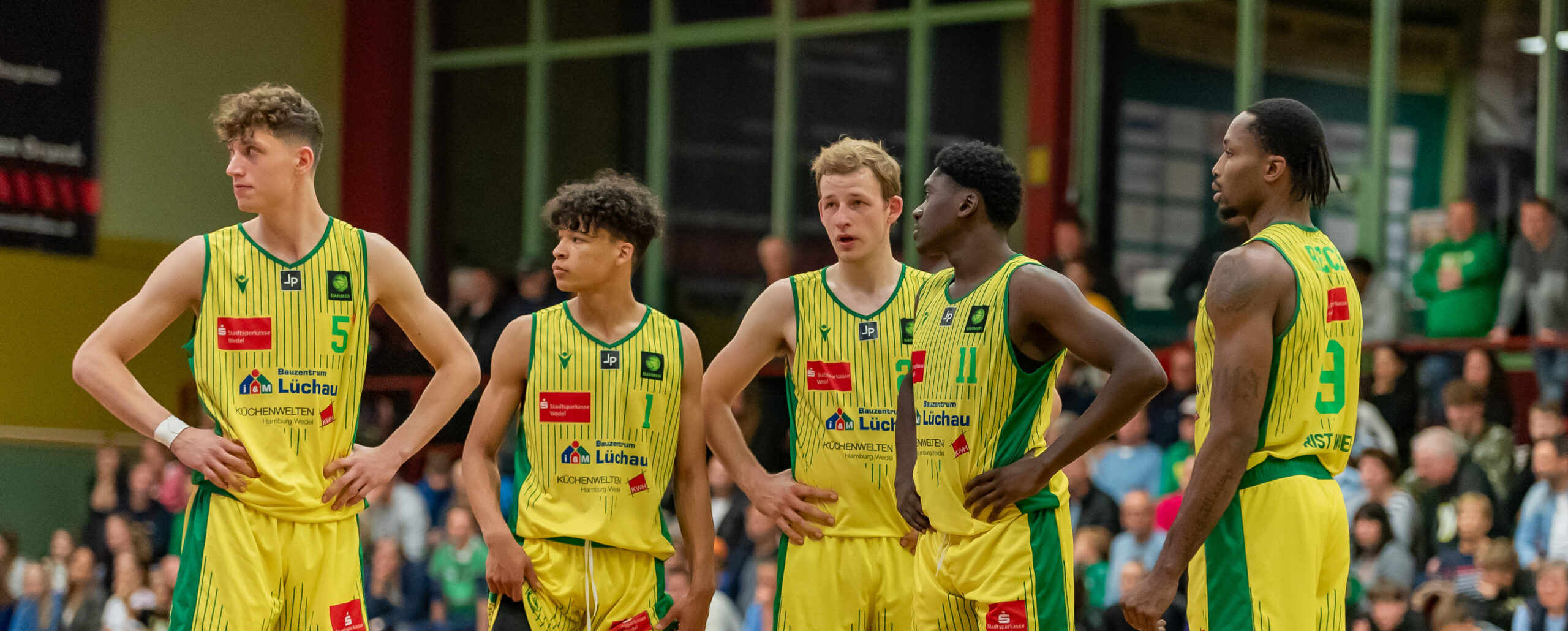 Fünf Basketballspieler in gelben Trikots mit grünen Details stehen nebeneinander auf dem Spielfeld während eines Spiels.