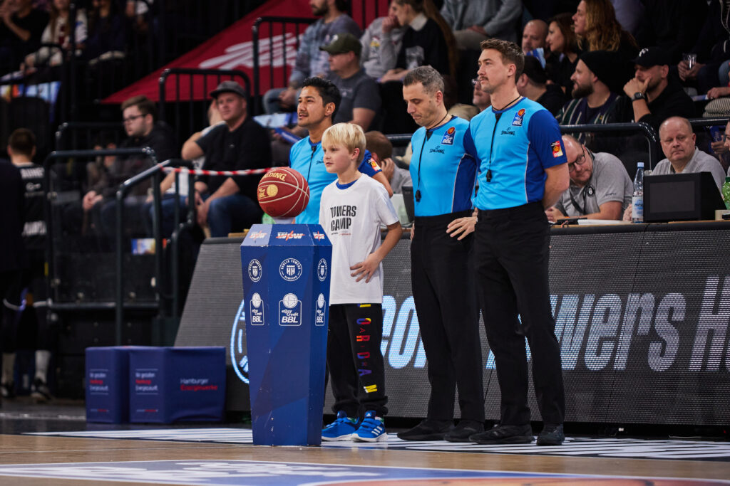 Tipoff-Kind posiert mit Schiedsrichter-Trio mit Ball und Stele im Vordergrund