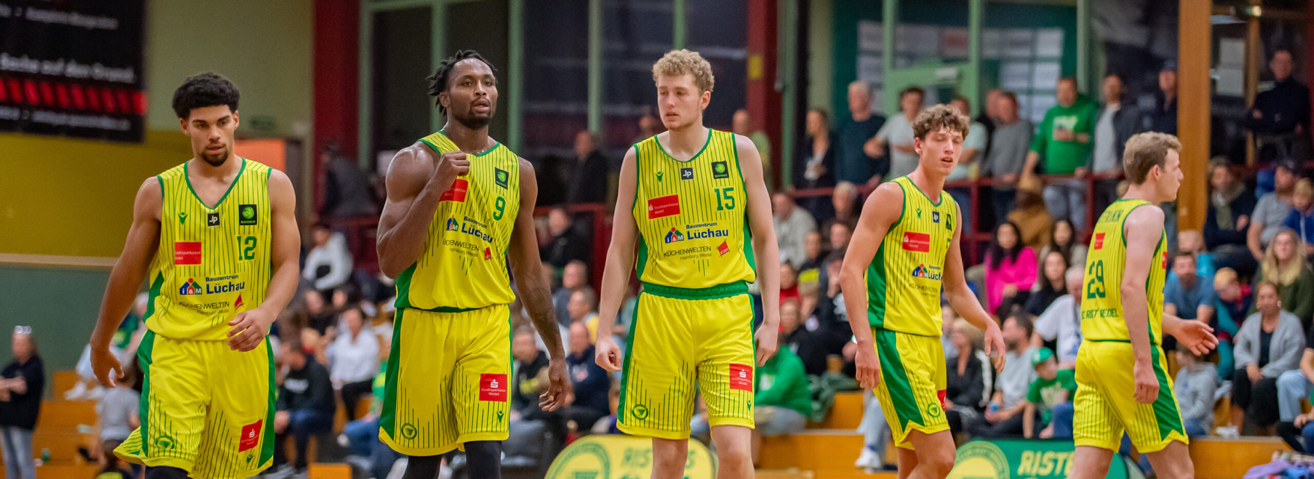 Fünf Spieler in gelb-grünen Uniformen stehen auf dem Spielfeld, im Hintergrund Zuschauer