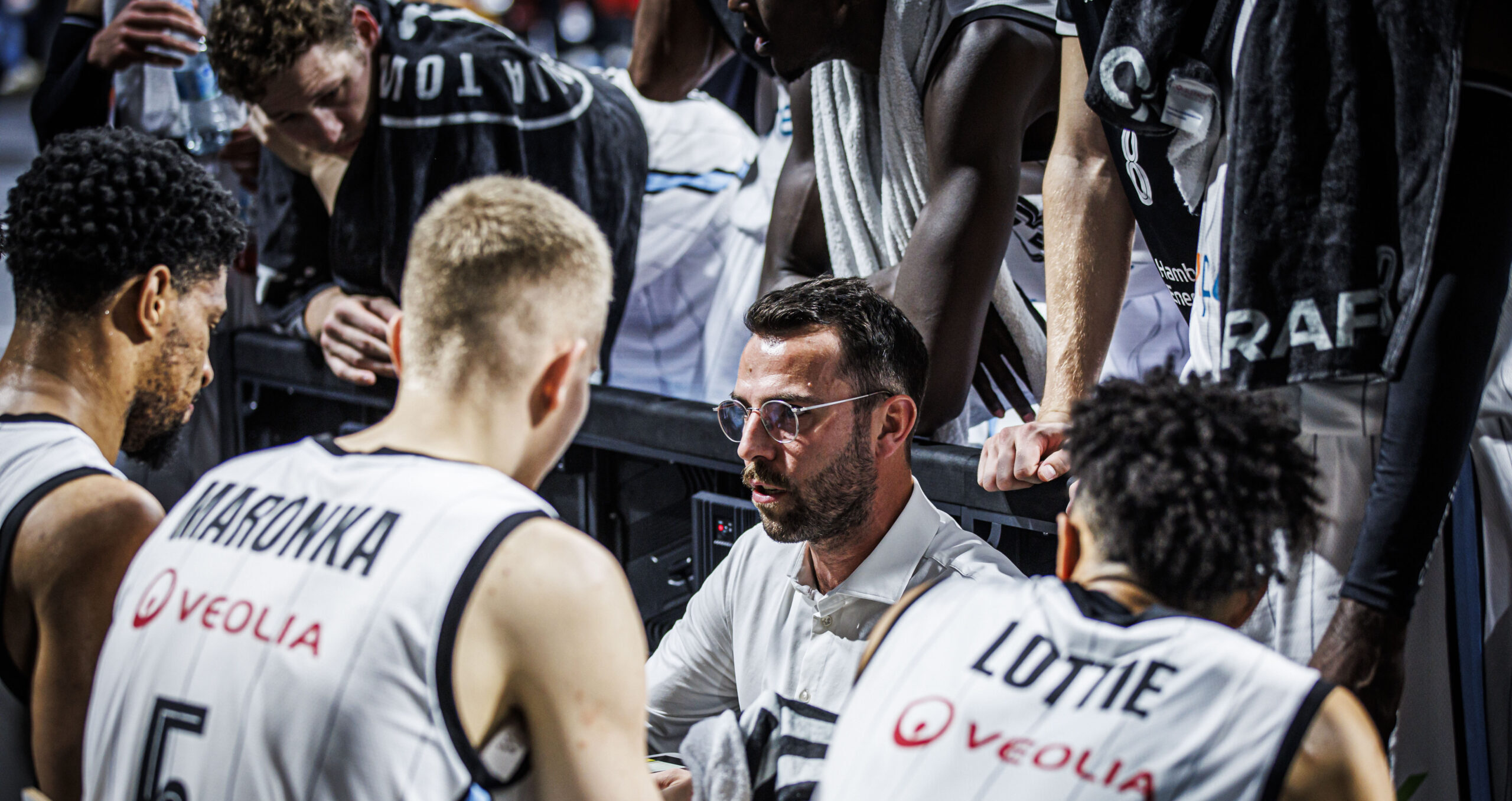 Basketballspieler Maronka und Lottie in weißen Trikots mit Veolia-Logo während einer Auszeit am Spielfeldrand