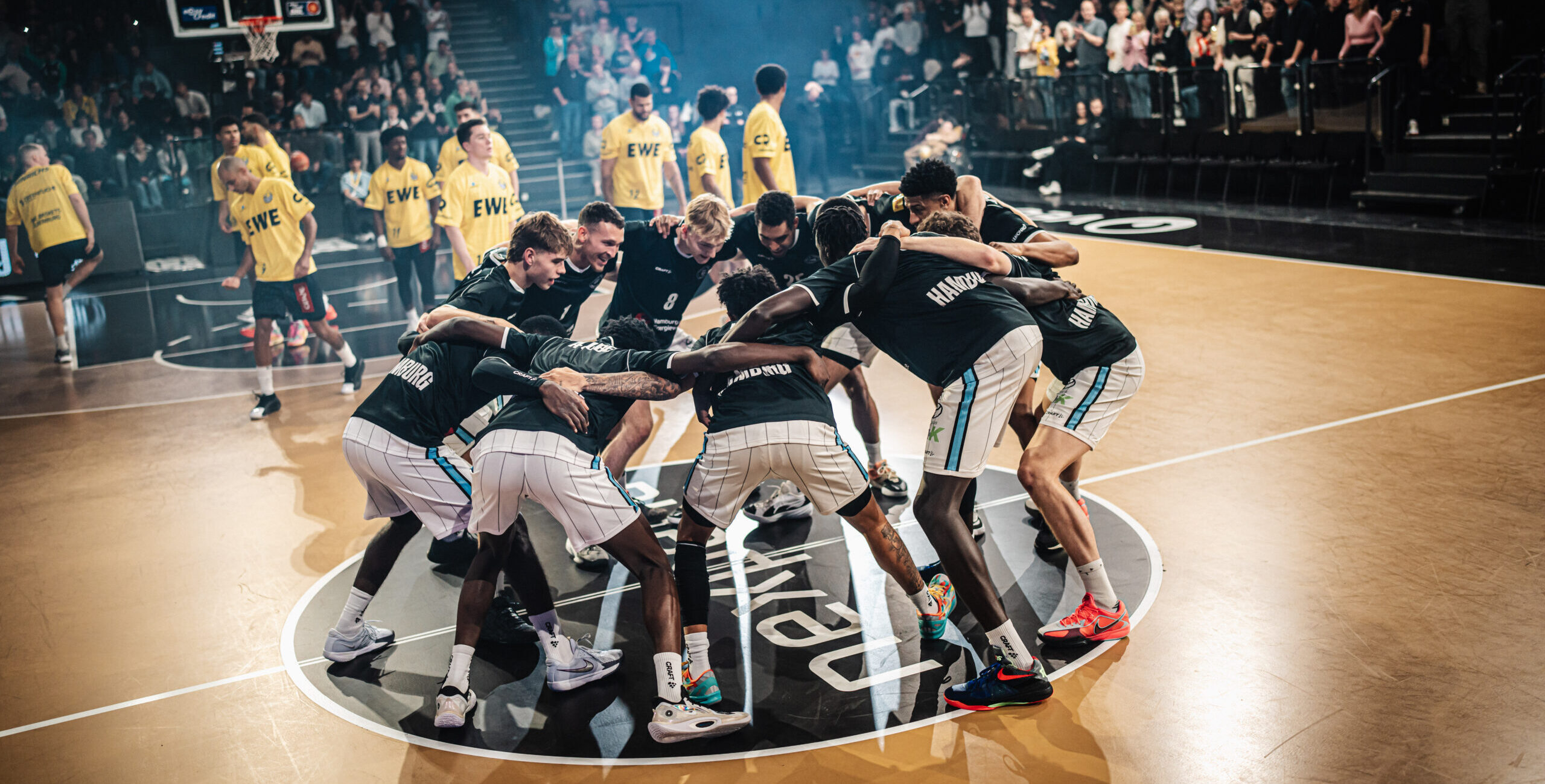 Basketballteam in schwarzen Trikots steht im Kreis auf dem Spielfeld und bereitet sich auf das Spiel vor