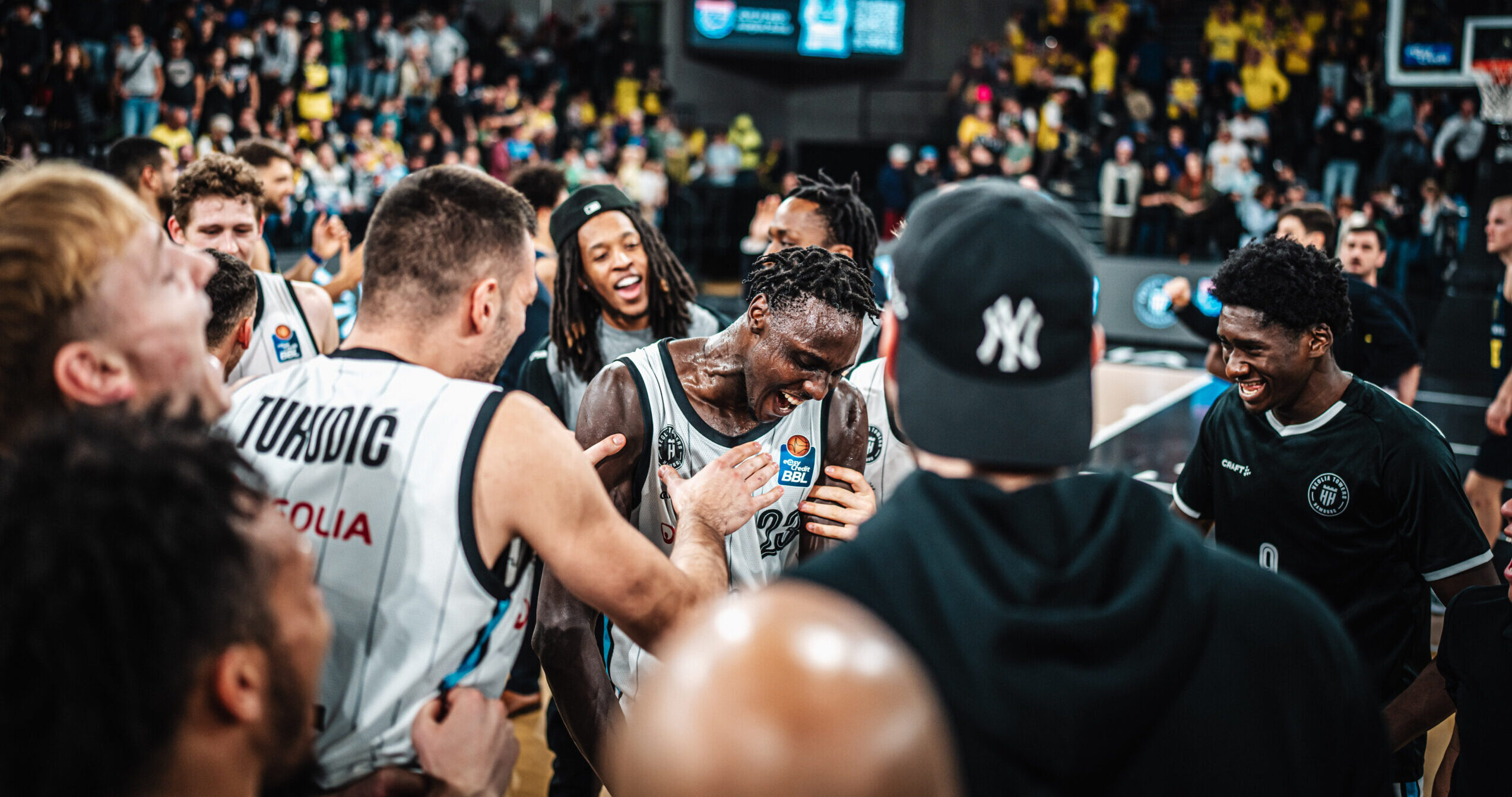 Basketballspieler in weißen Trikots in einer Spielpause, umgeben von Teammitgliedern und Zuschauern in einer Halle