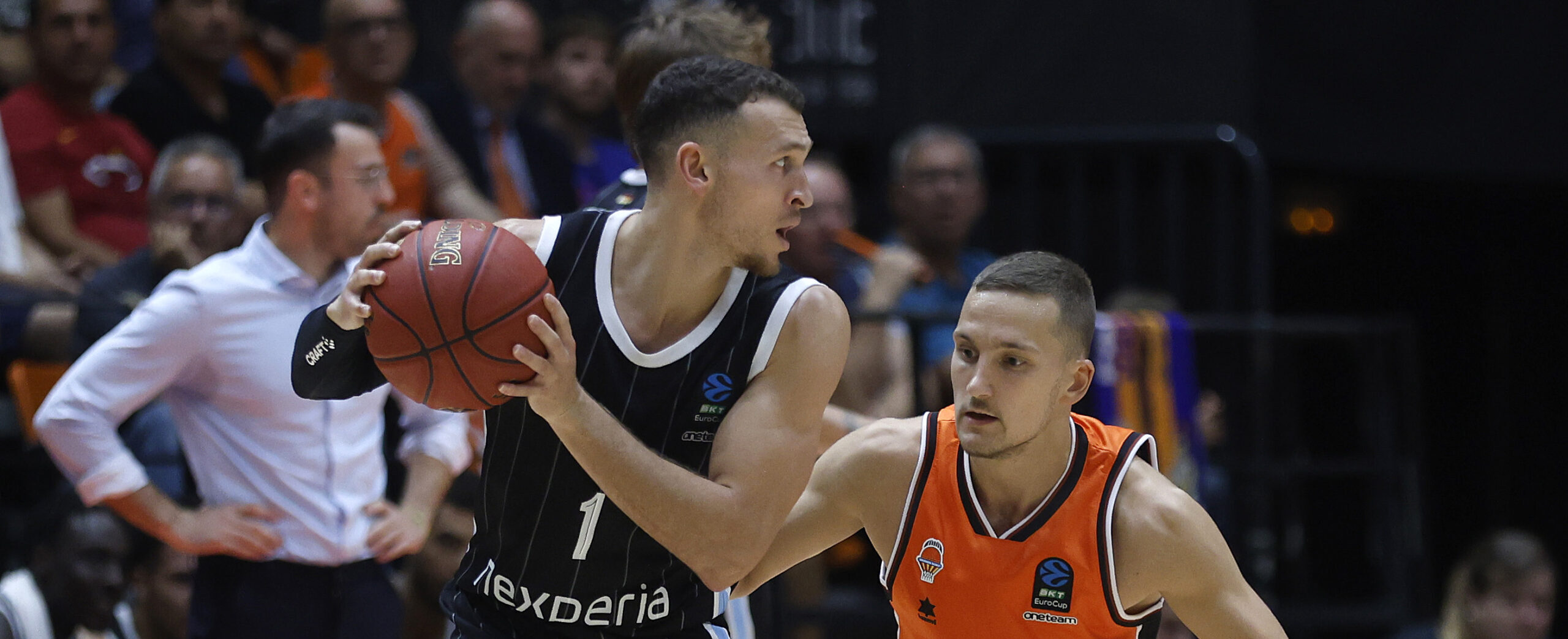 Basketballspieler in schwarzem Trikot mit Ball, verteidigt von Spieler in orangefarbenem Trikot während eines Spiels