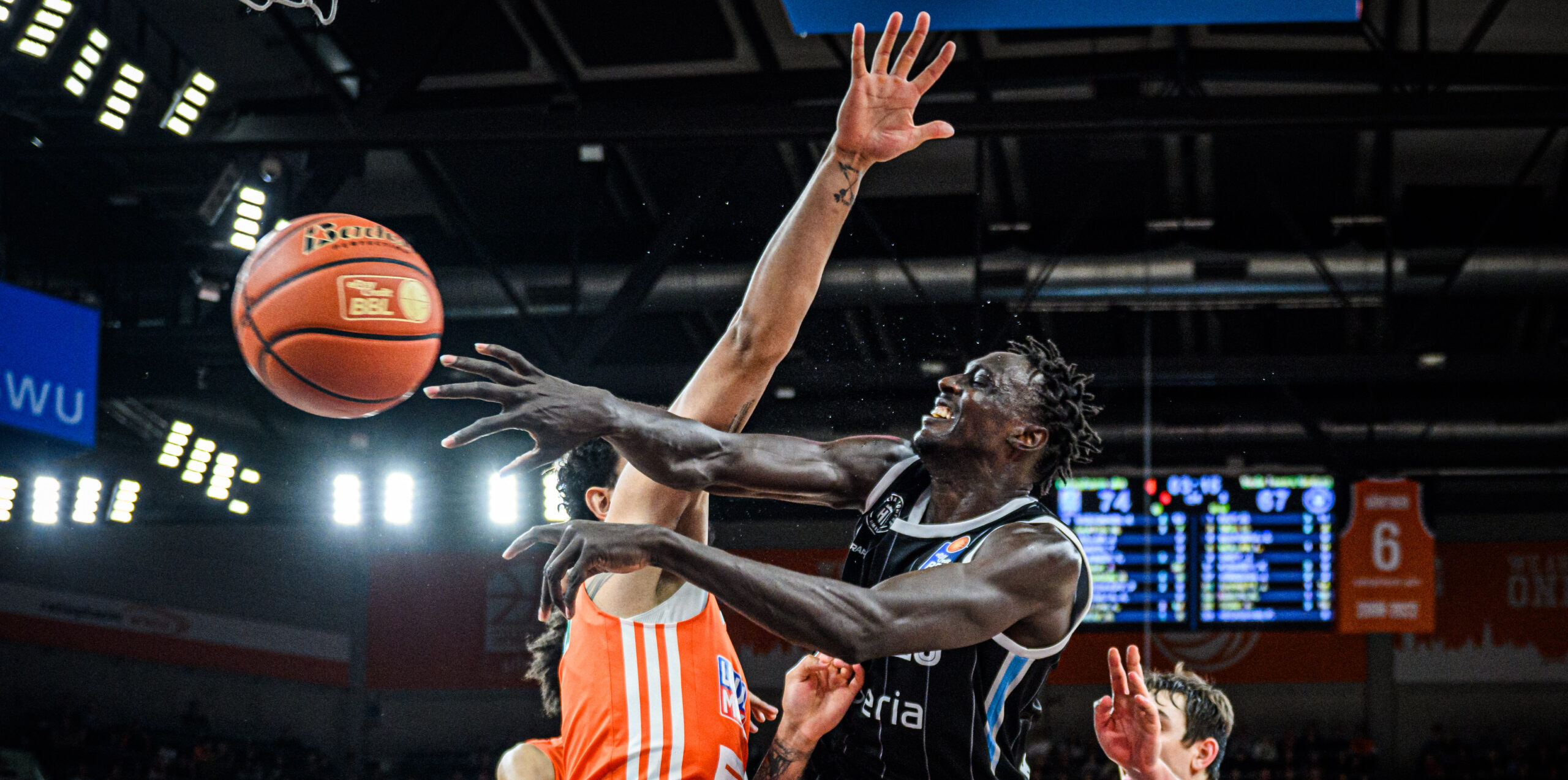 Basketballspieler in schwarzem Trikot wirft Ball, verteidigt von Spieler in orangefarbenem Trikot während eines Spiels