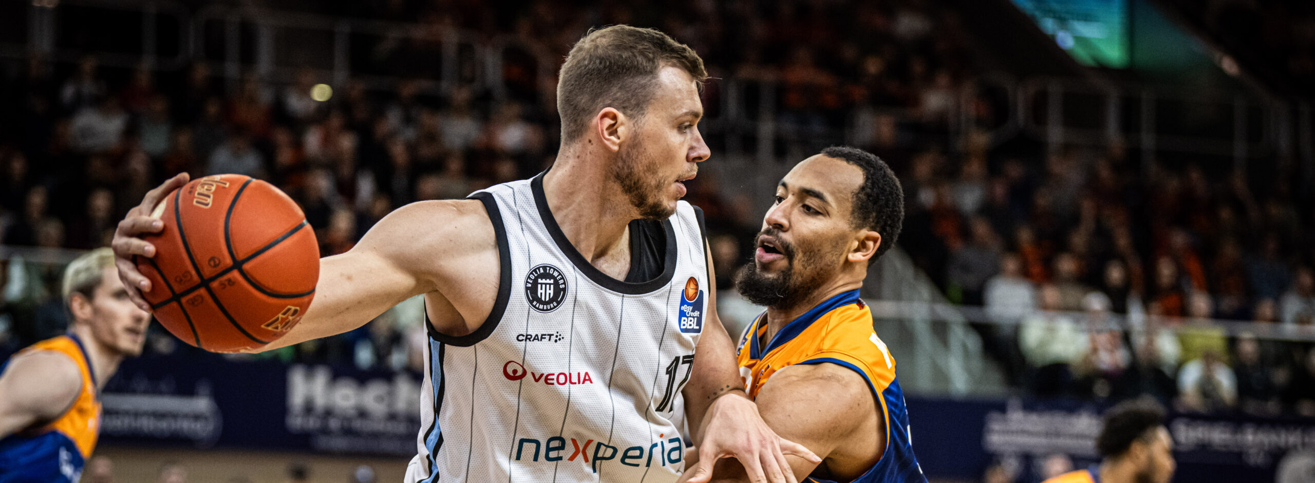 Niklas Wimberg in weißem Trikot mit Ball gegen aggressiv verteidigenden Spieler in orange-blauem Trikot