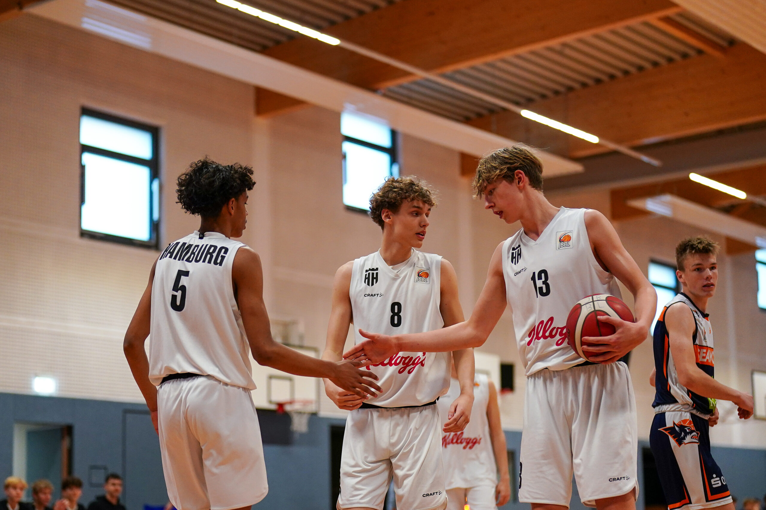drei junge Spieler in weißen Trikots klatschen bei einander ab, einer hält einen Basketball in der Hand