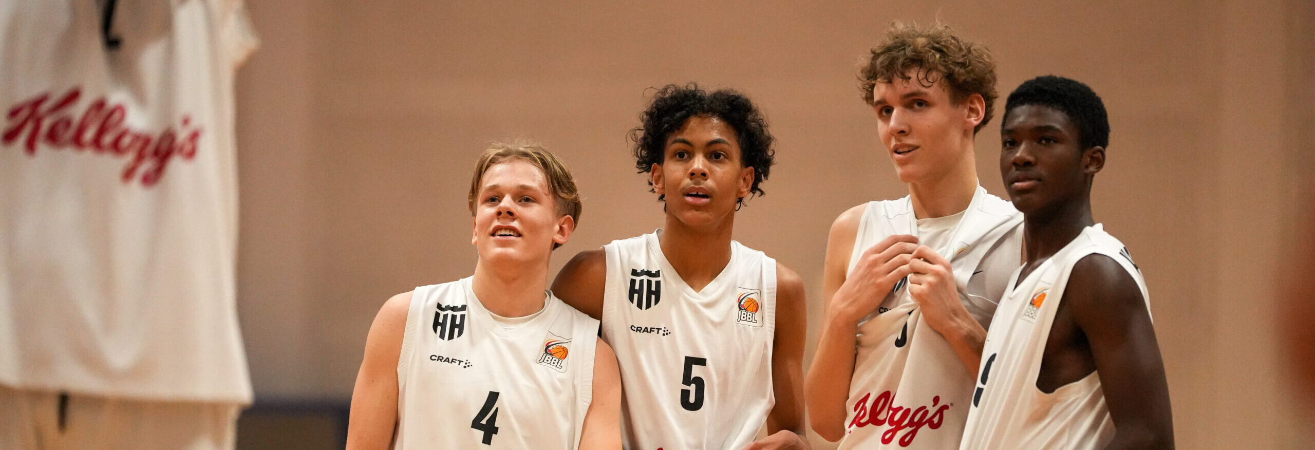 vier junge Basketballspieler in weißen Trikots stehen nebeneinander und blicken Richtung Spielfeld