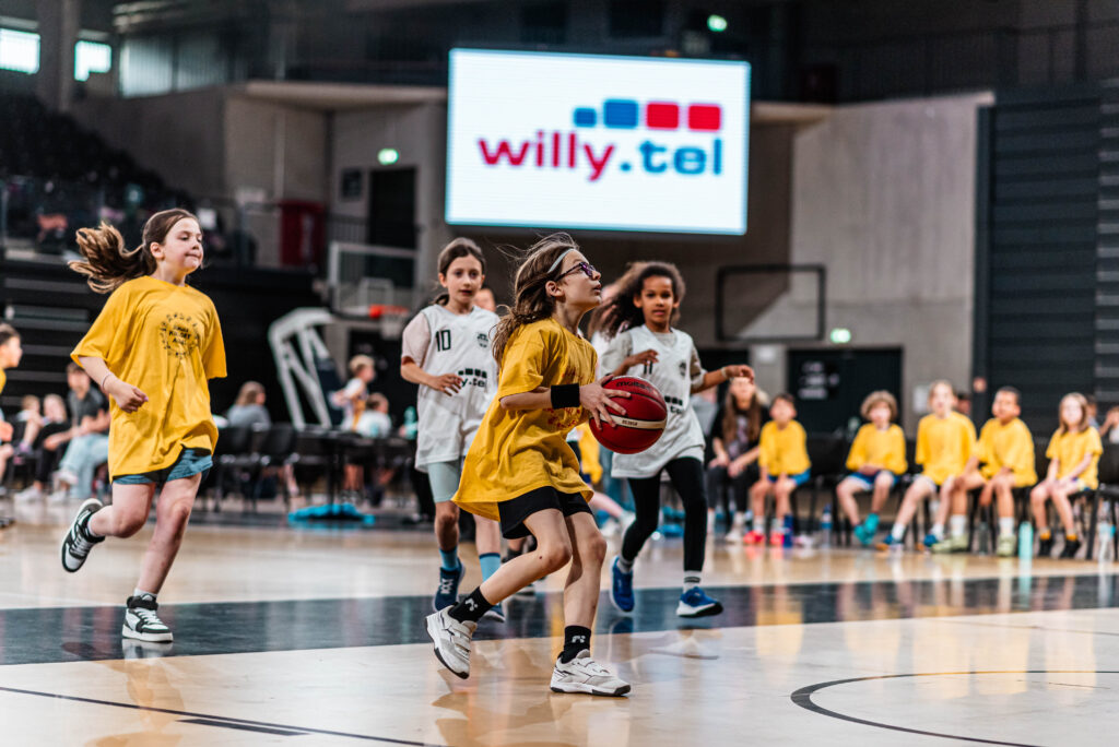 Kinder beim Basketball, im Hintergrund sitzen Kinder auf einem Stuhl, auf einer Wand steht willy.tel