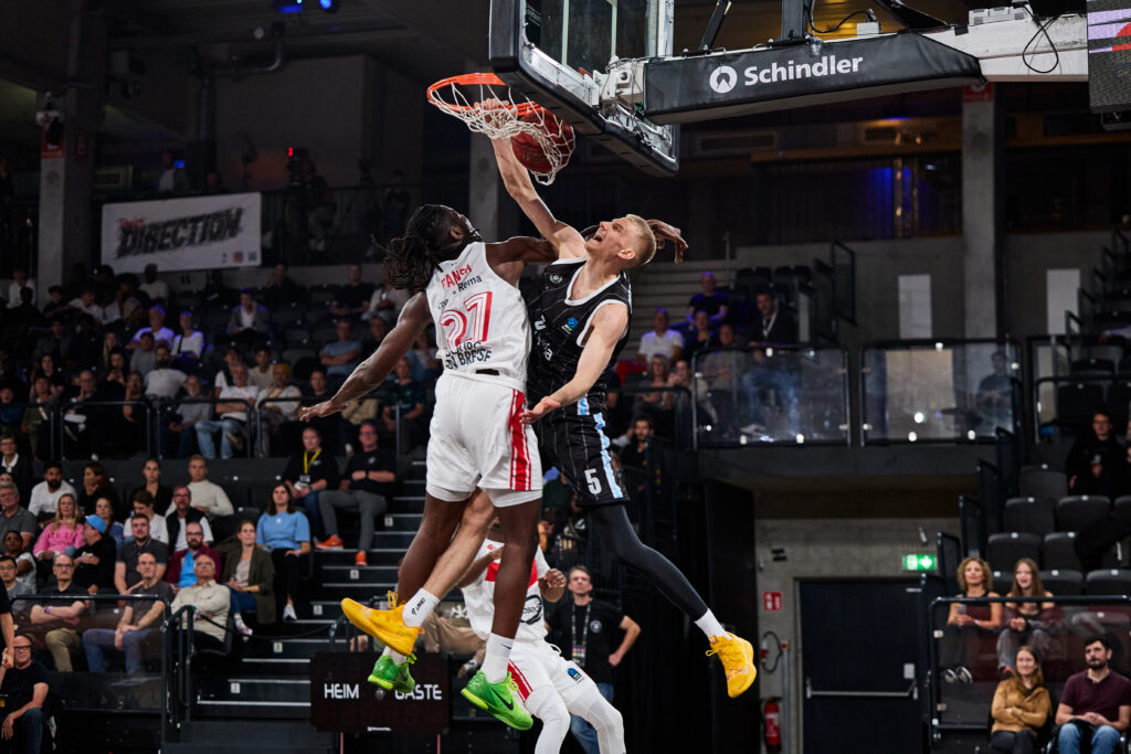 Spieler in schwarzem Trikot dunkt den Basketball über einen Spieler in weißem Trikot