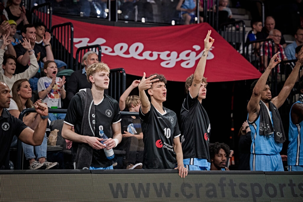 Basketball-Spieler in schwarzen T-Shirts feiern auf der Bank, im Hintergrund ein rotes Banner mit der Aufschrift Kellogg's