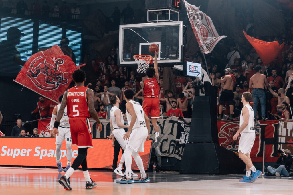 Aufnahme eines Basketballspiels zwischen Mannschaften mit roten und weißen Trikots. Ein Spieler im roten Trikto mit der Aufschrift Tokoto erzielt einen Korb. Im Hintergrund schwenken Fans Fahnen
