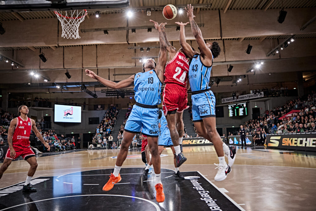 Basketballspieler in blauen und roten Trikots strecken sich nach dem Ball