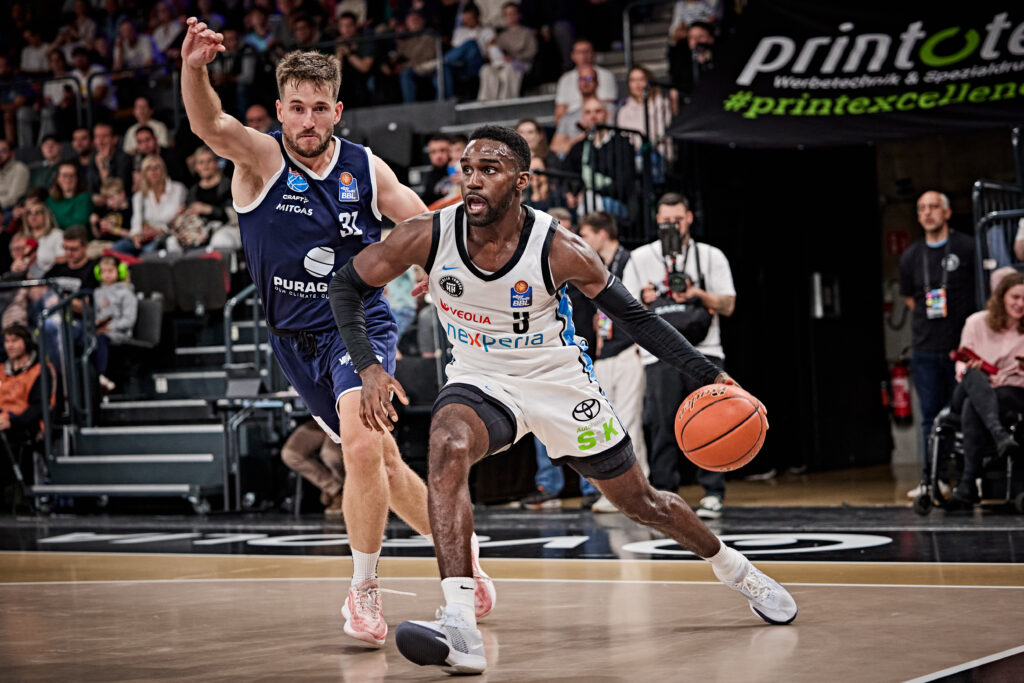 Basketballspieler in weißem Tritkot mit Ball in der Hand läuft an Gegenspieler in blauem Trikot vorbei