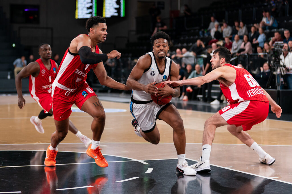 Basketballspieler mit weißem Trikot und Ball in der Hand wird von zwei Spielern in rotem Trikot verteidigt