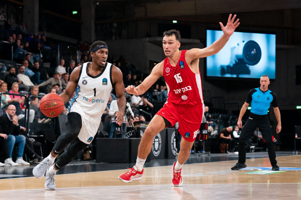 Basketballspieler mit weißem Trikot und Ball in der Hand wird von einemSpieler in rotem Trikot verteidigt