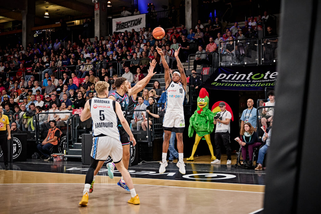 Basketballspieler in weißem Trikot beim Wurf in gefüllter Arena, im Hintergrund steht ein Huhn-Maskottchen
