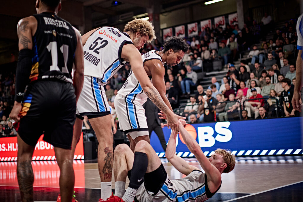 Zwei Basketballspieler in weißem Trikot helfen ihrem Mitspieler vom Boden hoch