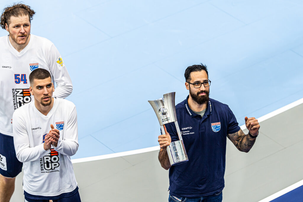 Spieler in T-Shirt bringt einen Pokal mit, reckt die Faust, daneben stehen zwei Spieler in Spielkleidung und klatschen