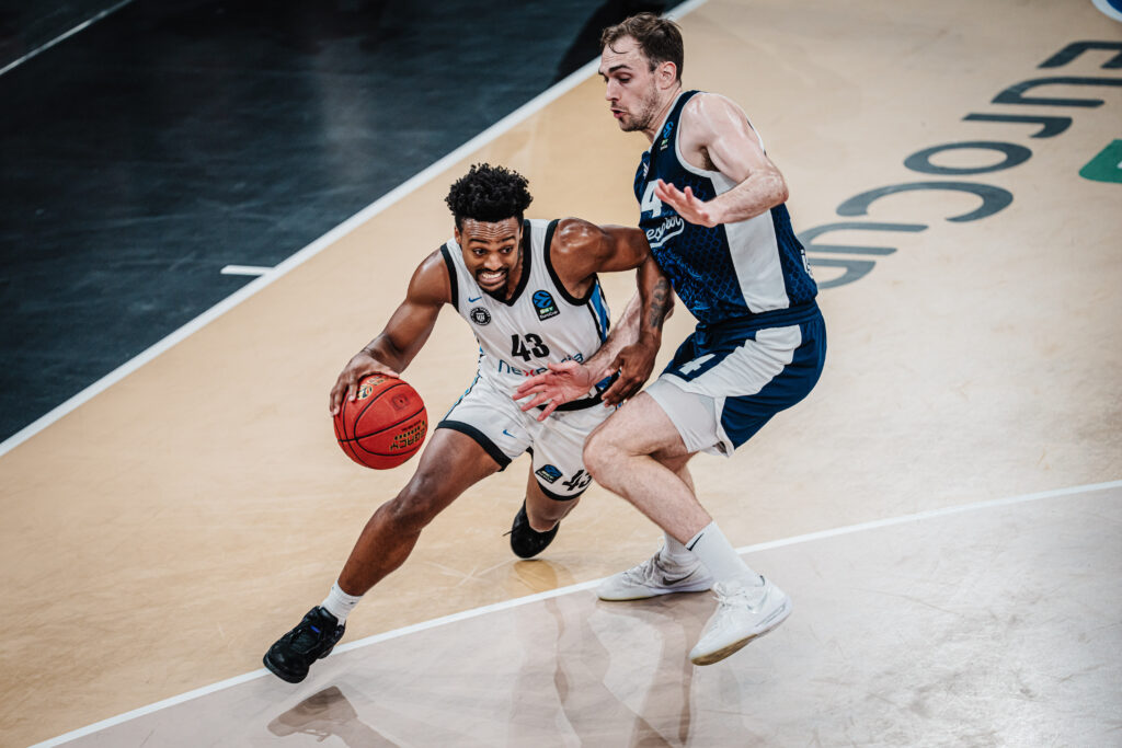 Basketballspieler in weißem Trikot mit Ball in der Hand zieht an Gegenspieler in blauem Trikot vorbei