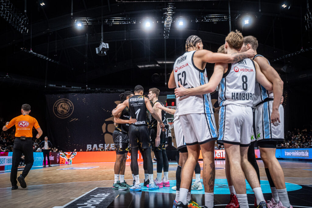 Mehrere Basketballspieler in weißen und schwarzen Trikots umarmen sich auf dem Spielfeld in einer Halle mit dunkler Decke und hellen Scheinwerfern.